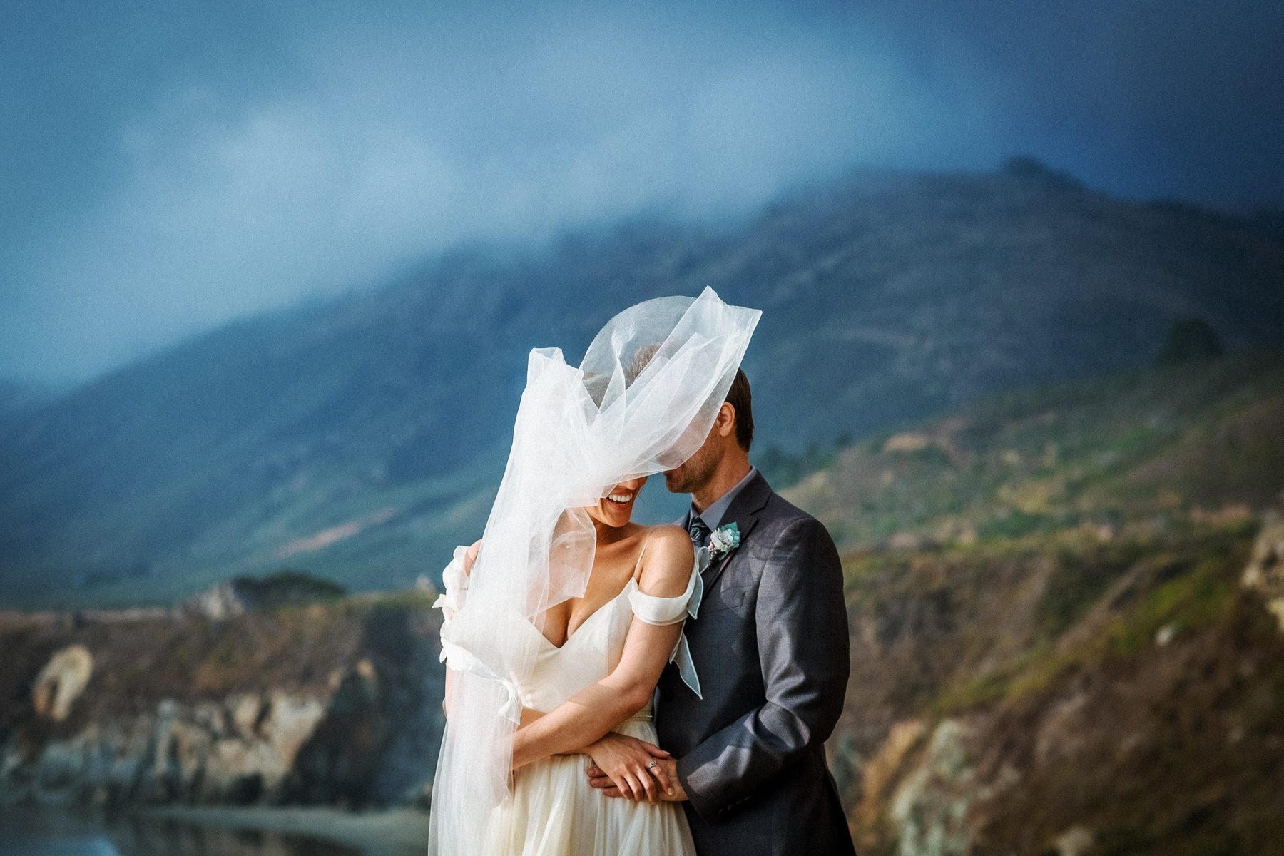 Bride and groom laughing beneath a windswept veil during their Big Sur wedding portraits